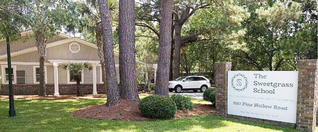 The Sweetgrass School building with welcoming entrance, surrounded by trees and landscaping, featuring a sign with the school's name and address.