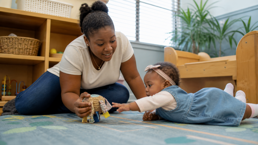 Caregiver engaging an infant with a soft toy on a classroom rug